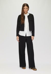Schwarze, cropped Jacke mit zwei vorderen Taschen, über einem weißen Hemd mit Knöpfen. Kombiniert mit lockeren schwarzen Hosen und braun akzentuierten Schuhen.