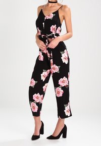 Combinaison noire à motif floral avec des fleurs roses, sans manches, bretelles ajustables, taille cintrée et pantalons courts ; portée avec des talons noirs.