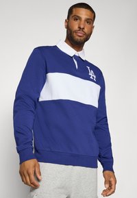 Blå sweatshirt med hvid vandret stribe og LA-logo på brystet. Har en krave-farve og ribbede manchetter. Blød stoftekstur.