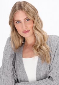 Šedý pletený cardigan s kabelkovou texturou, s hlubokým výstřihem do V a žebrovanými manžetami. Modelka má pod ním bílé tričko.