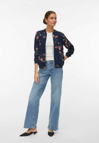 Kobieta stojąca z ręką na biodrze, ubrana w granatową kwiatową kurtkę bomberkę, biały top, szerokie niebieskie dżinsy oraz czarne buty na obcasie z ostrym noskiem.