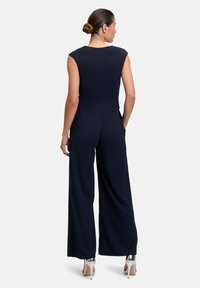 Combinaison bleu marine avec un corsage ajusté, des manches courtes et un design à jambes larges. Tissu lisse et détail de fermeture éclair dans le dos, portée avec des talons argentés.