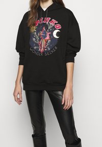 Svart huvtröja med en färgglad grafisk tryck av en fågel, stjärnor och måne med texten "WONDER SEEKER" nedanför. Mjuk material, oversized passform.