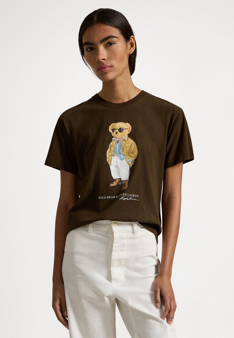 Brązowy bawełniany t-shirt z grafiką przedstawiającą niedźwiedzia w okularach przeciwsłonecznych i płaszczu. Ma okrągły dekolt i krótkie rękawy, zestawiony z białymi spodniami.