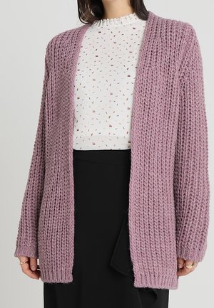 Kvinde iført en chunky strikket støvet lyserød cardigan over en hvid mønstret bluse og sort nederdel, stående mod en ensfarvet lys baggrund.