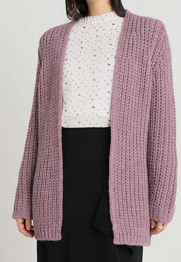 Femme portant un cardigan en grosse maille rose poudré sur un chemisier blanc à motifs et une jupe noire, debout devant un fond clair uni.