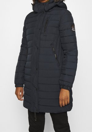 Person wearing a long, navy blue quilted winter coat with a hood, zippered chest and side pockets, and a logo patch on the left sleeve.