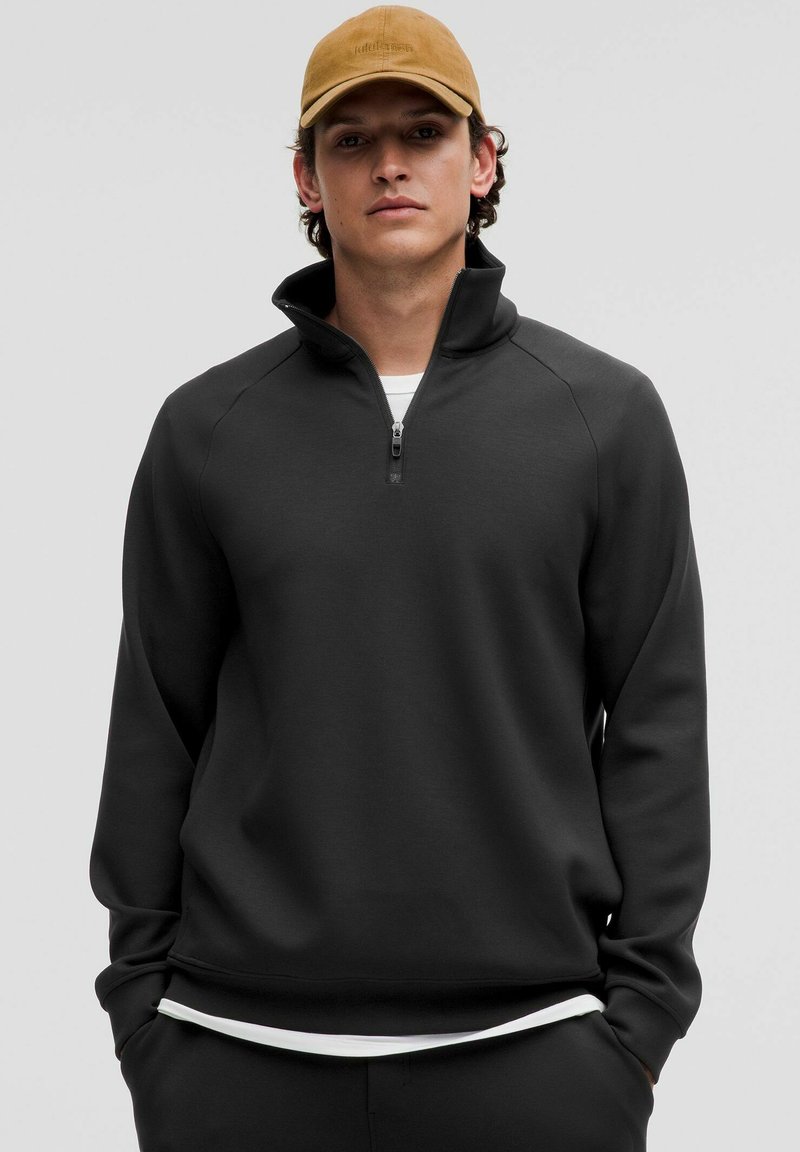 Sudadera negra con cremallera y cuello alto, y puños acanalados. Fabricada en un material suave y texturizado. Lleva una camiseta blanca debajo.