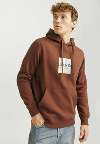 Barna kapucnis pulóver, elülső kenguru zsebből és egy fehér téglalap alakú logóból, amelyen zöld "JACK & JONES" felirat található.