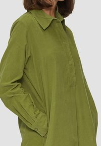 Camisa chaqueta de pana verde con un cuello puntiagudo, mangas largas y un corte relajado. Presenta una tapeta delantera y bolsillos laterales.