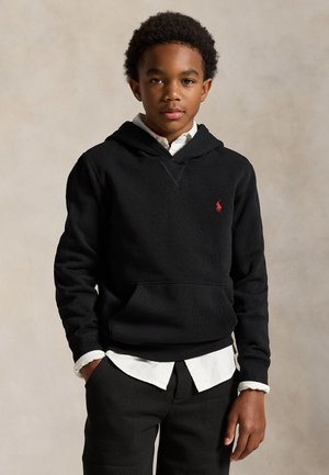 Garçon aux cheveux courts et bouclés portant un sweat à capuche noir par-dessus une chemise blanche à col, debout devant un fond beige uni.