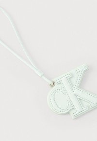Porte-clés en cuir blanc avec les initiales "CK" en relief cousues, attaché à une lanière bouclée sur un fond clair.