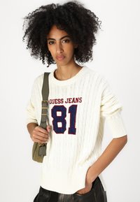 Maglione a maglia cable-knit color crema con il logo rosso e blu "Guess Jeans 81" sul davanti. Il modello tiene in mano una borsa a tracolla verde, indossando pantaloni neri.