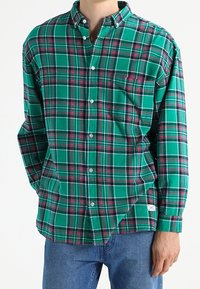 Camicia a quadri verdi con bottone, dotata di taschino sul petto, bottoni bianchi e colletto classico. Il materiale ha un aspetto morbido e testurizzato.