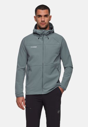 Homme portant une veste à capuche zippée grise et un pantalon noir, se tenant avec une main dans la poche devant un fond uni.