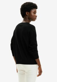 Maglione nero lavorato a maglia con dettagli testurizzati sulle spalle, polsini e orlo a coste. Indossato con jeans bianchi in una vista laterale.