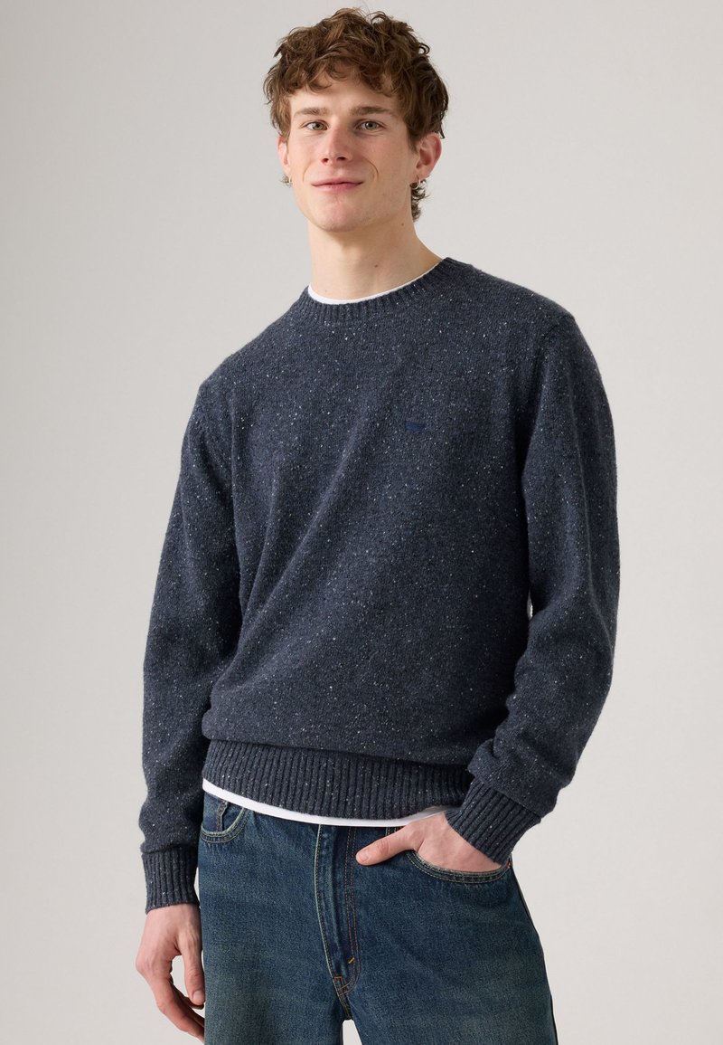 Marinblå prickig stickad tröja med ribbad nederdel och ärmslut, rund halsringning, bärs över en vit skjorta, ihop med blå jeans.