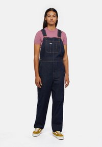 Denim overalls in dark blue with adjustable straps, front pocket, and gold stitching. Paired with a pink fitted shirt and yellow sneakers.