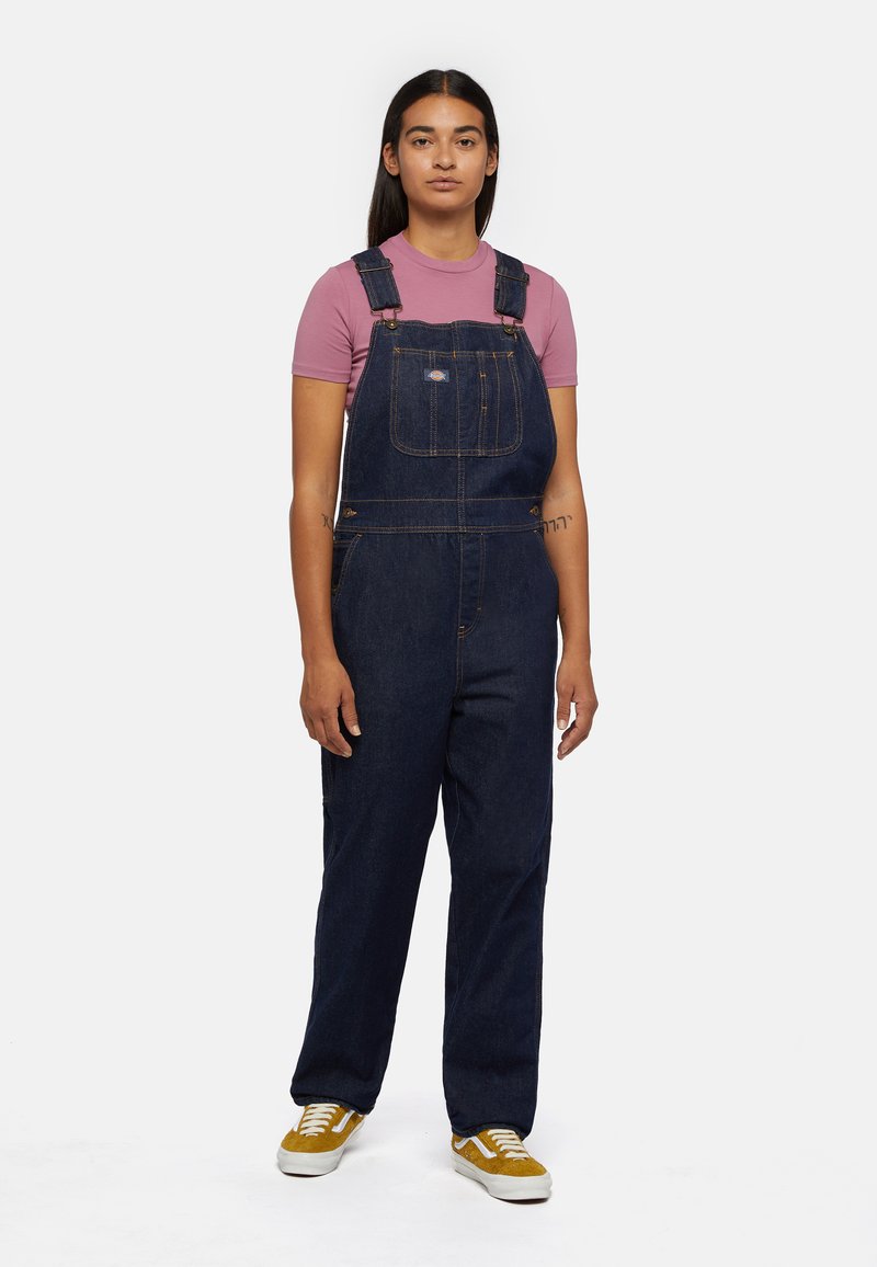 Denim overalls in dark blue with adjustable straps, front pocket, and gold stitching. Paired with a pink fitted shirt and yellow sneakers.