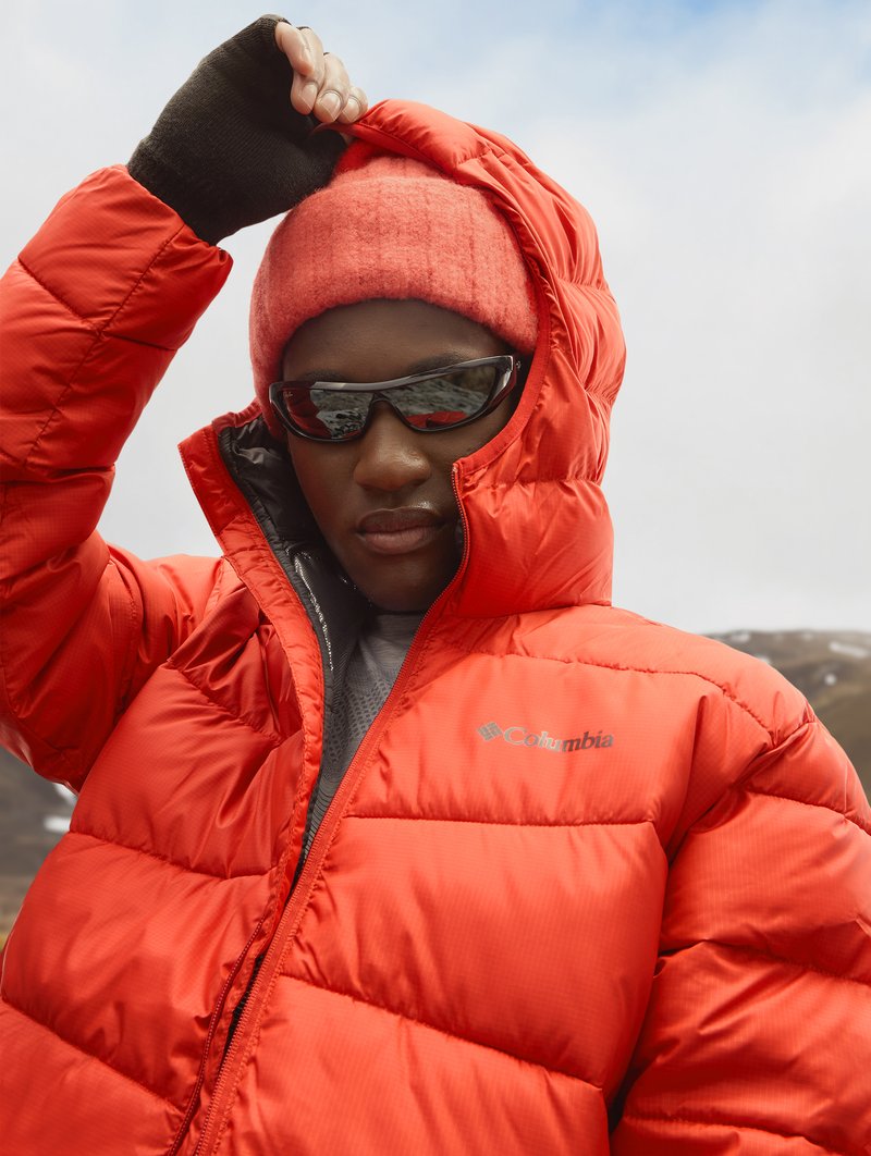Chaqueta acolchada roja con textura acolchada, con capucha y el logo de Columbia. El modelo lleva un gorro de punto rojo y gafas de sol.