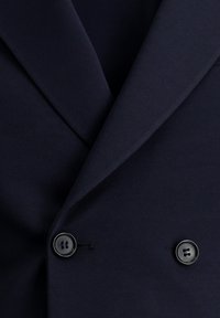 Primer plano de un blazer azul marino con tela texturizada, solapas afiladas y dos grandes botones negros. El diseño limpio presenta una apariencia ajustada.