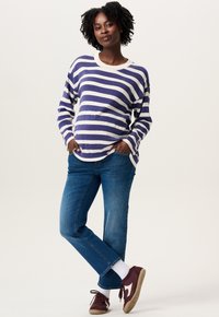 Femme avec des cheveux bouclés portant un pull rayé bleu et blanc, un jean bleu, des chaussettes blanches et des baskets bordeaux, se tenant avec les mains dans les poches.