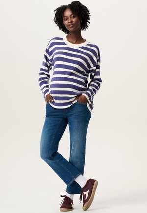 Vrouw met krullend haar die een blauwe en witte gestreepte trui, blauwe jeans, witte sokken en kastanjebruine sneakers draagt, terwijl ze met haar handen in haar zakken staat.