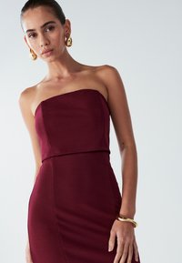 Burgundské bezramínkové šaty s přiléhavým střihem. Hladká tkanina, délka ke kolenům a horizontální šev. Zlatožluté kapkovité náušnice doplňují vzhled.