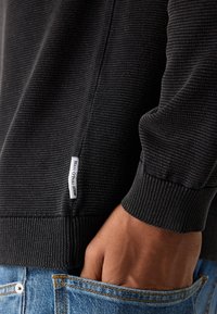 Main dans la poche portant un pull noir côtelé avec une étiquette "Marc O'Polo Denim" et un jean bleu avec des coutures visibles et un bouton.