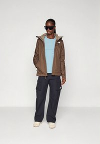 Brun vanntett jakke med lys grå fôr, blå t-skjorte, mørke cargobukser og beige sko. Modellen har på seg svarte solbriller.