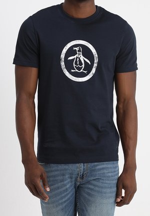 Mężczyzna w czarnej koszulce z białym logo pingwina w kółku, w połączeniu z niebieskimi dżinsami.