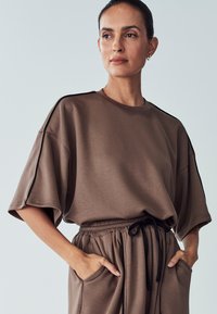 Brown oversized short-sleeve top with a rounded neckline, featuring contrasting black side accents and a relaxed fit. Soft fabric with pockets.