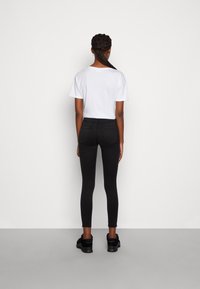 Mujer con cabello trenzado de pie de espaldas, vistiendo una camiseta blanca de manga corta, jeans ajustados negros y zapatillas negras sobre un fondo liso.