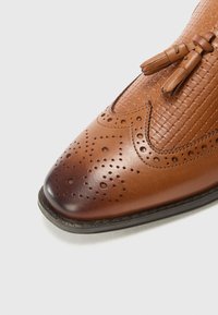 Zapato de vestir de cuero marrón con detalles perforados, parte superior texturizada y punta afilada. Presenta borlas de cuero en los cordones.