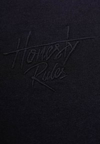 Mustekankaan musta neulekangas, jossa kohotettu, brodeerattu teksti "Honesty Rules" kauniissa kalvossa, taustalla sileä tekstuuri.
