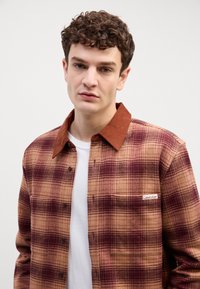 Smešani smešak smeđa i burgundska karirana košulja s okovratnikom od kotona, jednim džepom na prsima i oznakom logotipa Calvin Klein na džepu.