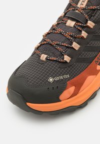 Czarno-pomarańczowe buty do biegania po szlakach z teksturowaną siateczkową cholewką, logo GORE-TEX oraz wytrzymałą gumową podeszwą. Posiada wielobarwne sznurowadła i wzór siatki.