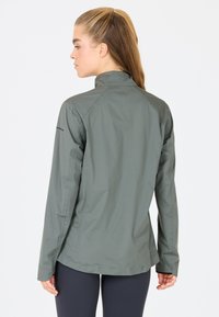 Lichte, grijsgroene jas met een hoge kraag en lange mouwen. Heeft een soepele, waterafstotende stof en een getailleerd ontwerp.