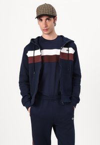 Sweatshirt azul-marinho com fecho, com riscas brancas e castanhas, combinado com uma t-shirt azul-marinho de gola redonda e calças de fato a condizer. Boné com padrão pied-de-poule.