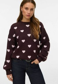 Pull bordeaux avec motifs de cœurs rose clair, poignets côtelés et encolure ronde, associé à un jean bleu foncé et une ceinture noire.