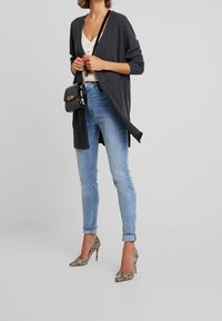 Lång, mörkgrå ribbade kofta med öppen framsida, bärs över en ljus topp, kombinerad med smala ljusblå jeans och klackar med ormskinnsmönster. Svart handväska som accent.