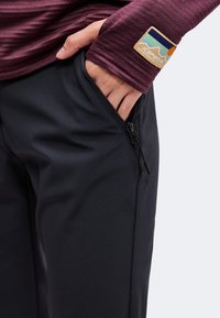 Main dans la poche zippée d'un pantalon noir, portant une chemise bordeaux à manches longues avec un patch montagne et soleil au poignet.