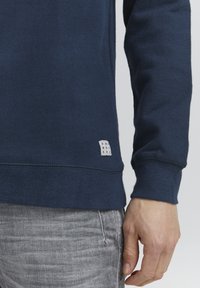 Felpa blu scuro con una finitura testurizzata, caratterizzata da un piccolo etichetta bianca con il testo "ONLY WORKERS" vicino all'orlo e polsini a costine.