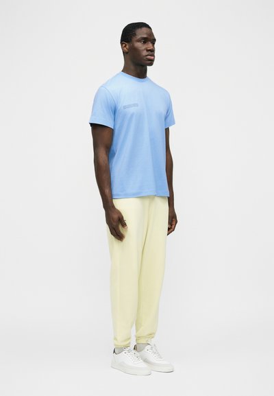 Άνδρας που στέκεται πλάγια φοράει γαλάζιο μπλουζάκι, κρεμ παντελόνι φόρμας και λευκά αθλητικά παπούτσια μπροστά σε έναν απλό ανοιχτόχρωμο φόντο.