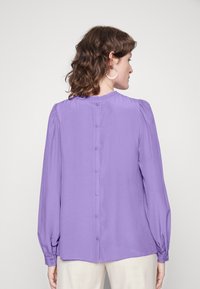 Blusa lilás de manga longa com decote franzido, fecho com botão nas costas e bainha arredondada; tecido suave e leve.