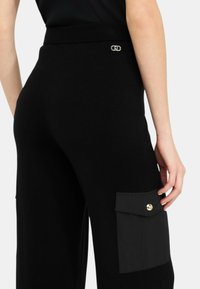 Schwarze weit geschnittene Hose mit strukturierter Oberfläche, ausgestattet mit einer großen Seitentasche mit Knopfverschluss und einem elastischen Bund. Details aus goldfarbenem Metall.