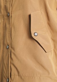 Light brown jacket featuring a textured fabric with snap buttons and a side pocket. Smooth finish with a relaxed fit.