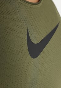 Top deportivo de color verde oliva, hecho de una tela texturizada, con un prominente logo en negro. El diseño incluye un escote redondo y una textura de malla transpirable.