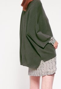 Överdimensionerad olivgrön ribbad tröja med vida ärmar, lager på lager över en mönstrad vit och svart klänning med geometriskt mönster.