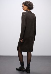 Dunkelbraunes Strickkleid mit hohem Kragen, langen Ärmeln, seitlichen Schlitzungen und gerippten Akzenten, kombiniert mit schwarzen Knöchelstiefeln.